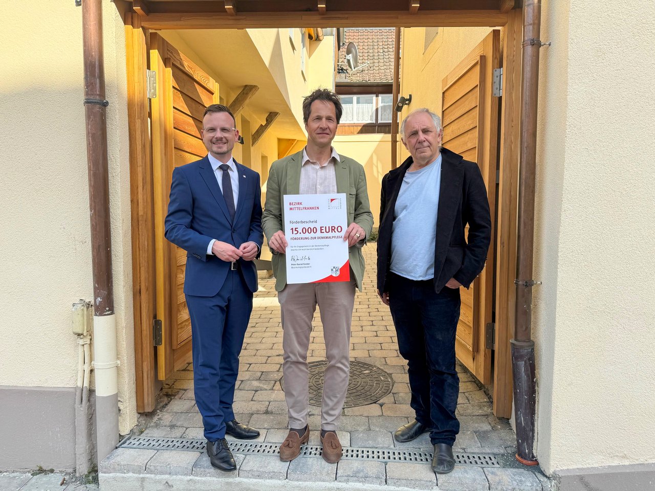 Hermann von Rotenhan nimmt in Erlangen den Förderbescheid über 15.000 Euro von Bezirkstagspräsident Peter Daniel Forster entgegen. Zusammen mit einem weiteren Mann stehen alle drei für das Foto im Türrahmen des Hauseingangs des sanierten Gebäudes und halten den Förderbescheid in die Kamera.