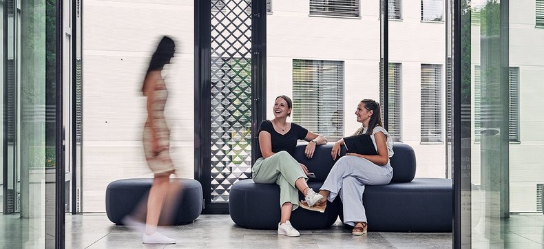 Blick durch die Eingangstür des Bezirksrathauses. Es sitzen zwei Frauen auf einer Sitzgelegenheit und begrüßen eine Dame, die gerade vorbeiläuft.