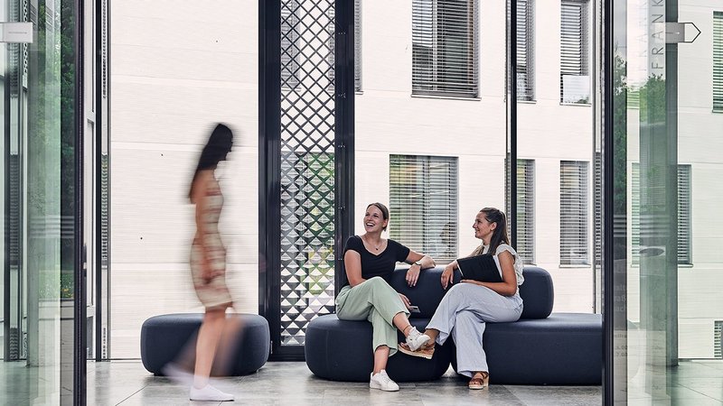 Blick durch die Eingangstür des Bezirksrathauses. Es sitzen zwei Frauen auf einer Sitzgelegenheit und begrüßen eine Dame, die gerade vorbeiläuft.