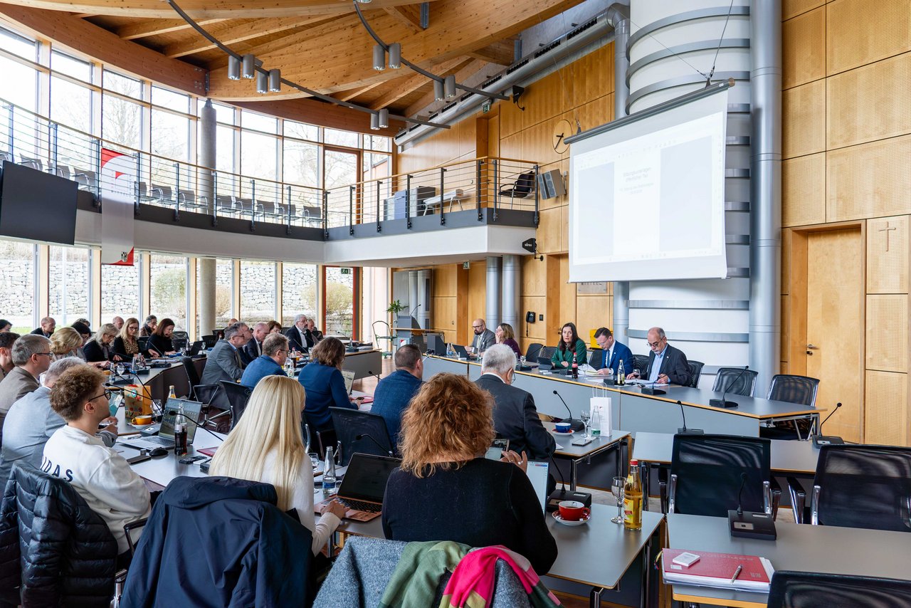 Bild aus dem voll besetzten Sitzungssaal: Die Bezirksrätinnen und Bezirksräte sitzen an ihrem Platz und hören Bezirkstagspräsident Peter Daniel Forster zu, der vorne am Präsidiumstisch sitzt.