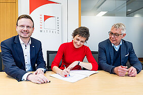 Drei Personen sitzen am Tisch zur Vertragsunterzeichnung