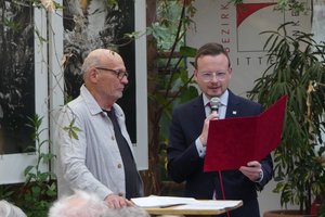 Bezirkstagspräsident Peter Daniel Forster verliest die Urkunde an Günter Derleth, der links neben ihm an einem Stehtisch steht.
