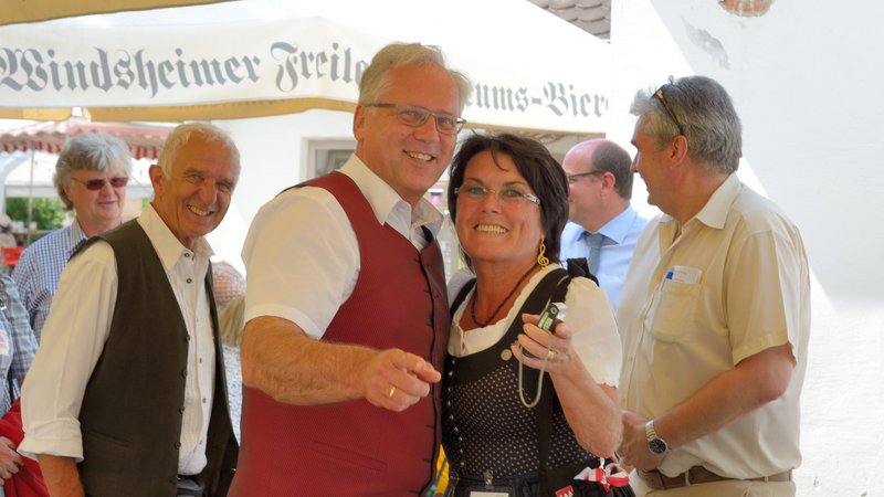 Zwei Menschen in Tracht lachen und zeigen in die Kamera. Im Hintergrund lachen stehen weitere Menschen und lachen in die Kamera.