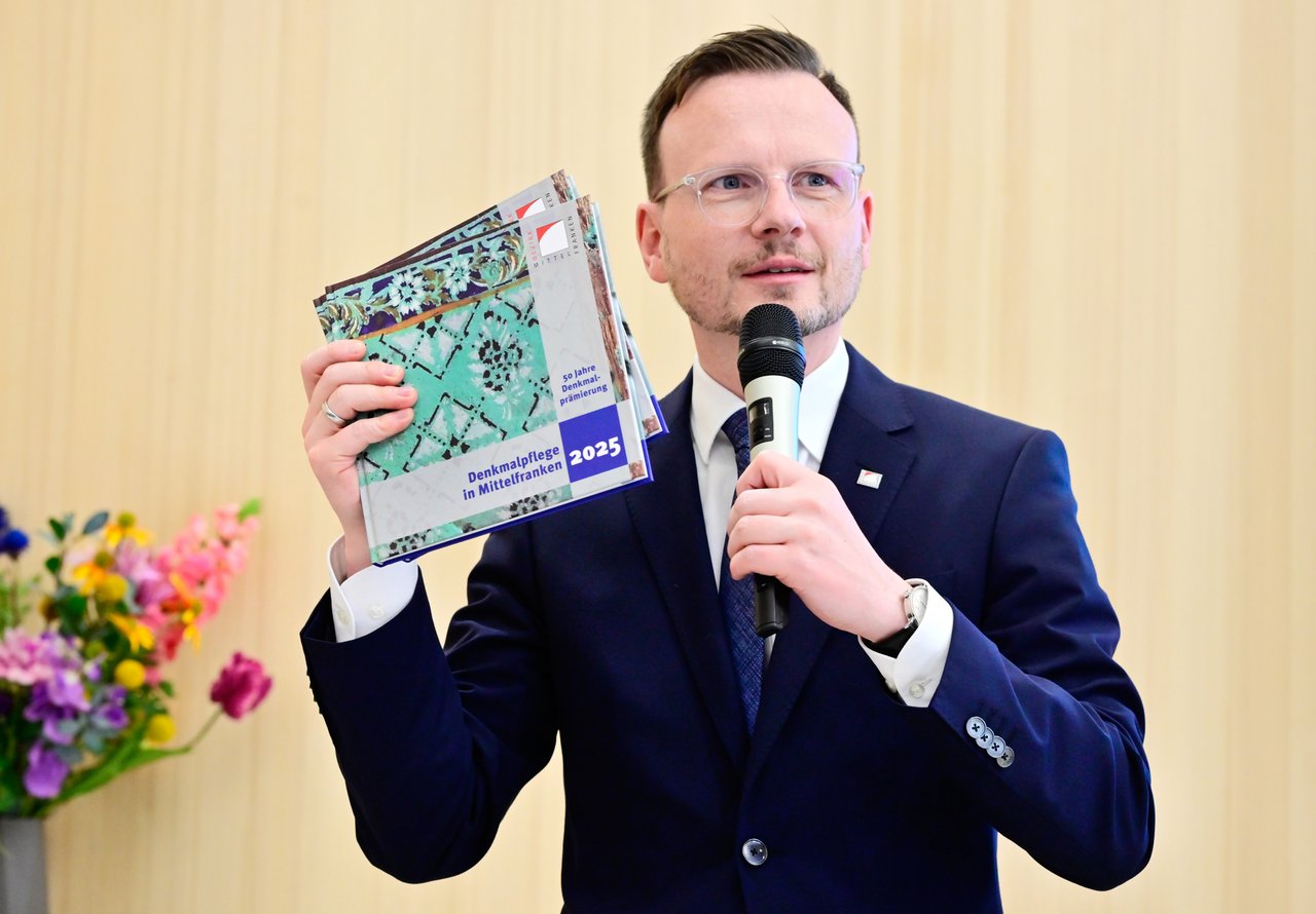 Bezirkstagspräsident Peter Daniel Forster spricht in ein Mikrofon zu den Gästen und hält dabei den Begleitband zur Denkmalprämierung 2025 in der Hand nach oben.