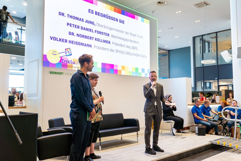 Bezirkstagspräsident Peter Daniel Forster steht auf einer Bühne und spricht in ein Mikrofon. Im Hintergrund ist ein großes LED-Display zu sehen.