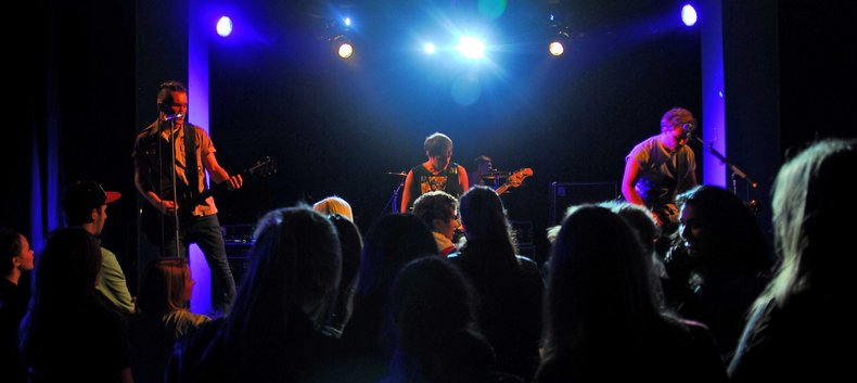 Blick auf eine Bühne, auf der gerade eine Band auftritt. Davor sind in der Dunkelheit die Silhouetten einiger Menschen aus dem Publikum zu sehen.