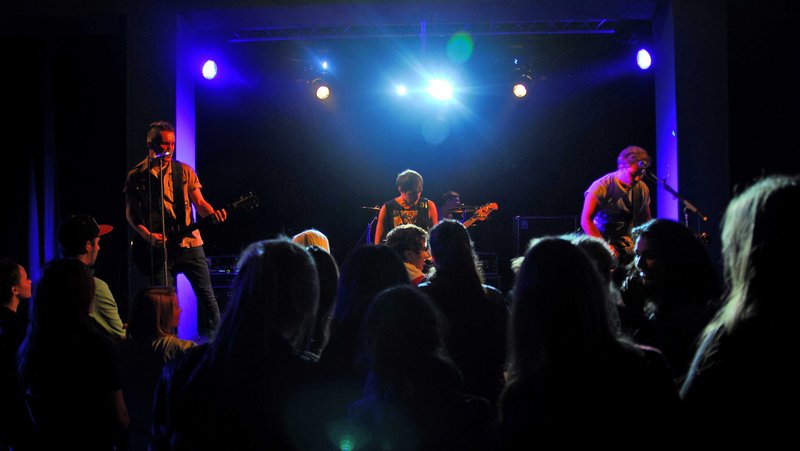 Blick auf eine Bühne, auf der gerade eine Band auftritt. Davor sind in der Dunkelheit die Silhouetten einiger Menschen aus dem Publikum zu sehen.