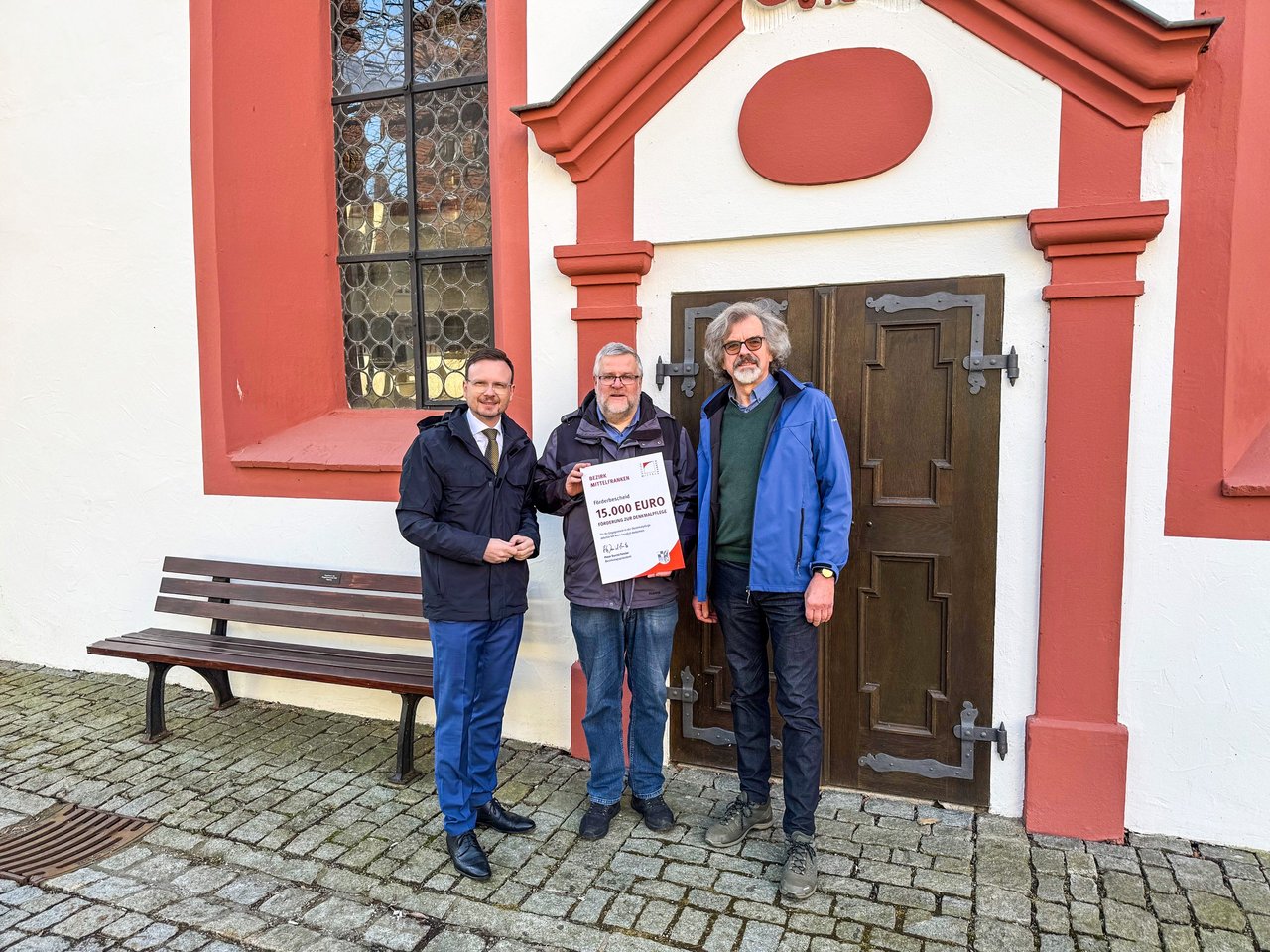 Die drei beteiligten Personen stehen außen vor der Kirche und halten den Förderbescheid hoch.