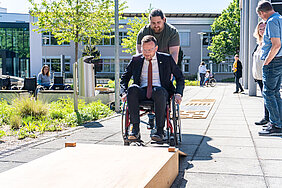Bezirkstagspräsident Peter Daniel Forster sitzt in einem Rollstuhl und versucht gerade, im Rollstuhlparcours auf eine Rampe hinaufzufahren. Dabei wird er von einem Mann unterstützt, der hinter ihm den Rollstuhl hält und leicht mitschiebt.