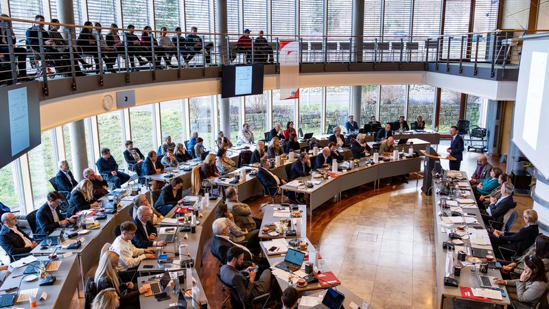 Der Sitzungssaal von der Empore fotografiert. Es läuft eine Sitzung. Der Saal ist voll besetzt.