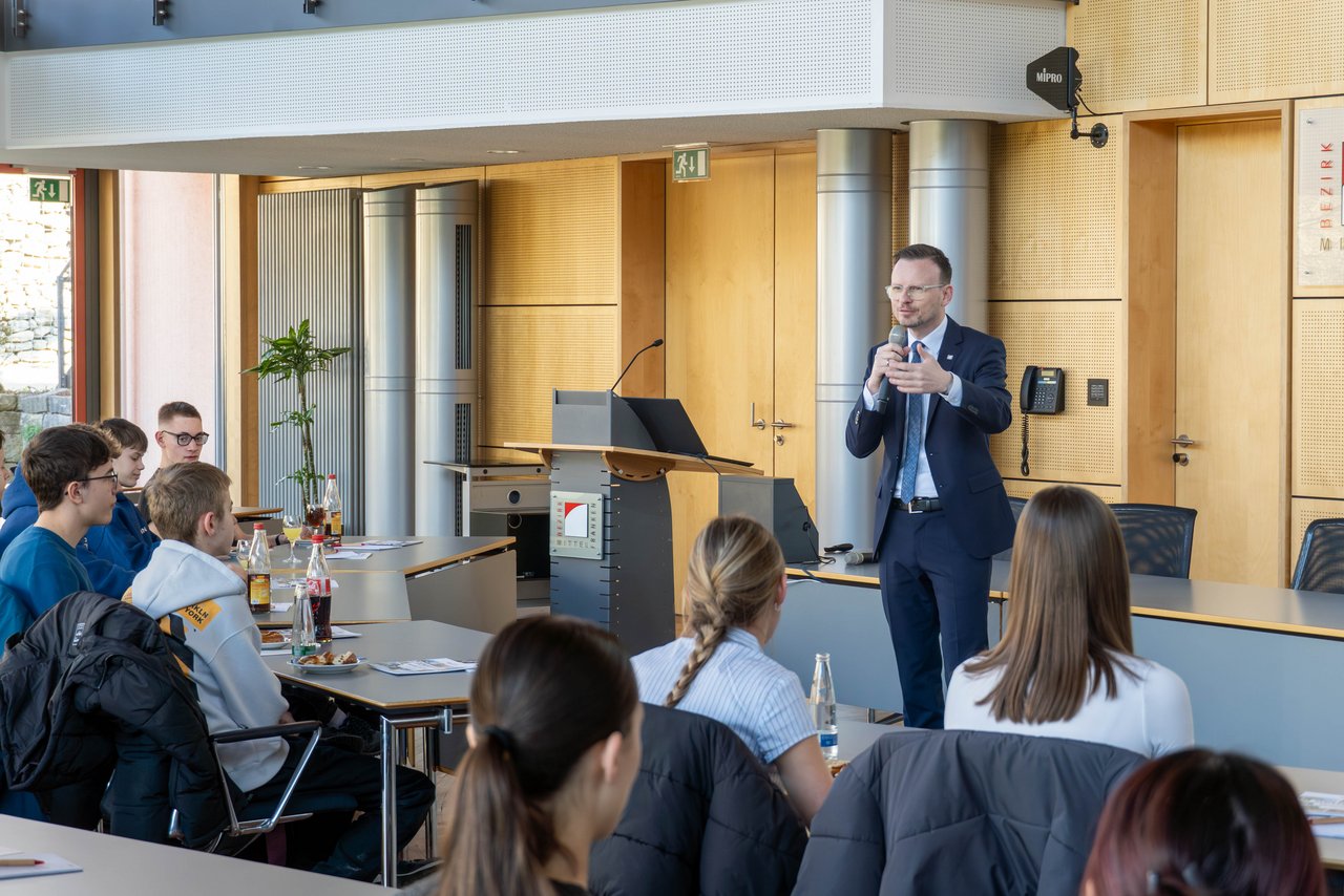 Bezirkstagspräsident Peter Daniel Forster steht mit dem Mikrofon in der Hand vor dem Plenum und spricht zu den Schülern.