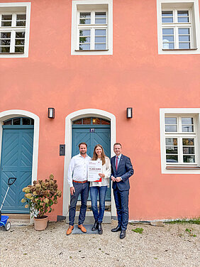 Drei Menschen stehen vor einem Haus mit einem Scheck in den Händen.