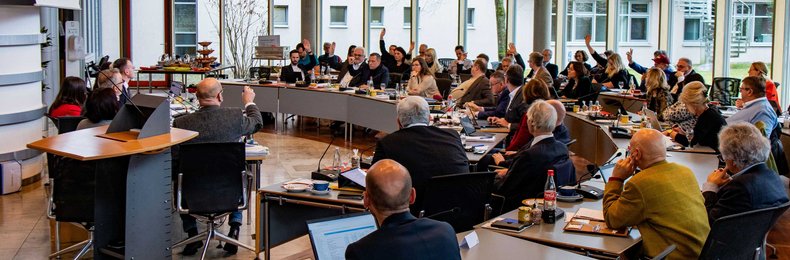 Blick in den vollen Sitzungssaal des Bezirksrathauses während einer Sitzung. Viele Abgeordnete des Bezirkstags heben ihre Hand, da gerade eine Abstimmung läuft.