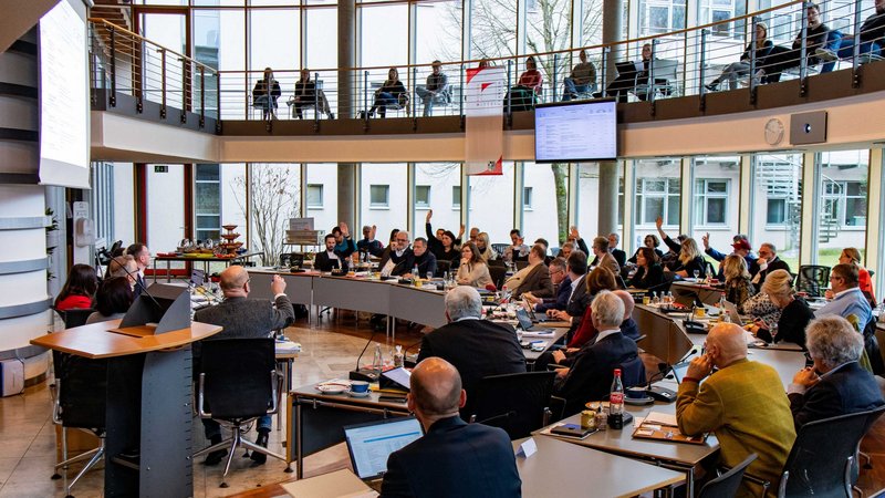 Blick in den vollen Sitzungssaal des Bezirksrathauses während einer Sitzung. Viele Abgeordnete des Bezirkstags heben ihre Hand, da gerade eine Abstimmung läuft.