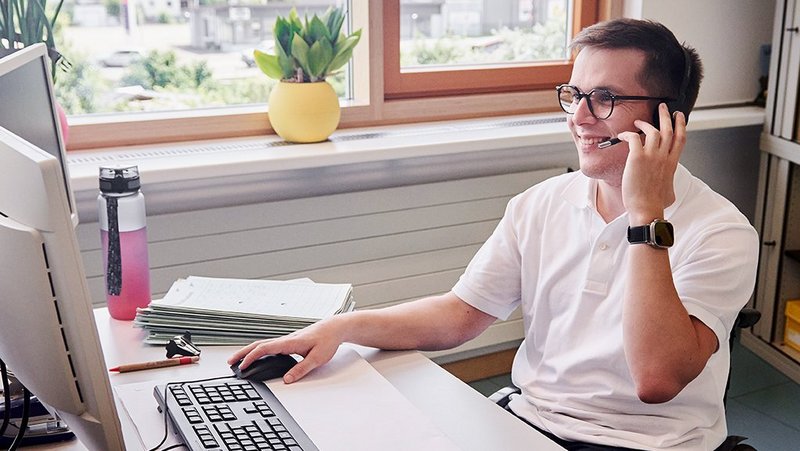 Ein Mann im Rollstuhl sitzt an seinem Schreibtisch, schaut auf seinen PC-Bildschirm und telefoniert mit seinem Headset.