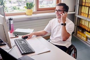 Ein Mann im Rollstuhl sitzt an seinem Schreibtisch, schaut auf seinen PC-Bildschirm und telefoniert mit seinem Headset.