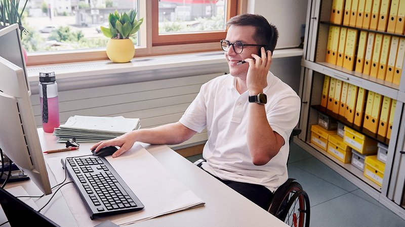 Ein Mann im Rollstuhl sitzt an seinem Schreibtisch, schaut auf seinen PC-Bildschirm und telefoniert mit seinem Headset.