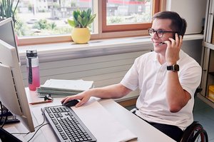Ein Mann im Rollstuhl sitzt an seinem Schreibtisch, schaut auf seinen PC-Bildschirm und telefoniert mit seinem Headset.
