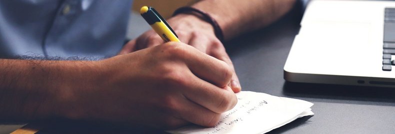 Eine Person sitzt an einem Schreibtisch und schreibt mit einem Kugelschreiber etwas auf einen Zettel. Man sieht nur die Hände und Arme der Person.