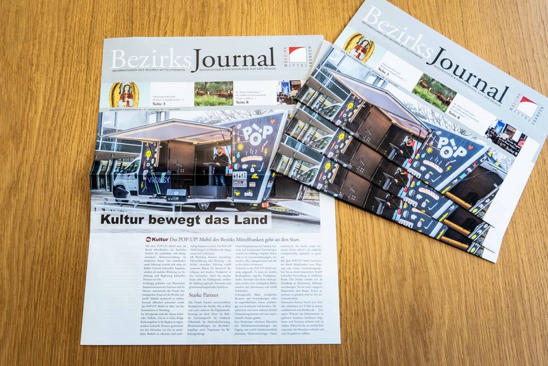 Mehrere Ausgaben der aktuellen Ausgabe des Bezirks-Journals liegen aufeinander auf einem Tisch. Daneben liegt eine Ausgabe, von der die Titelseite aufgeschlagen ist.