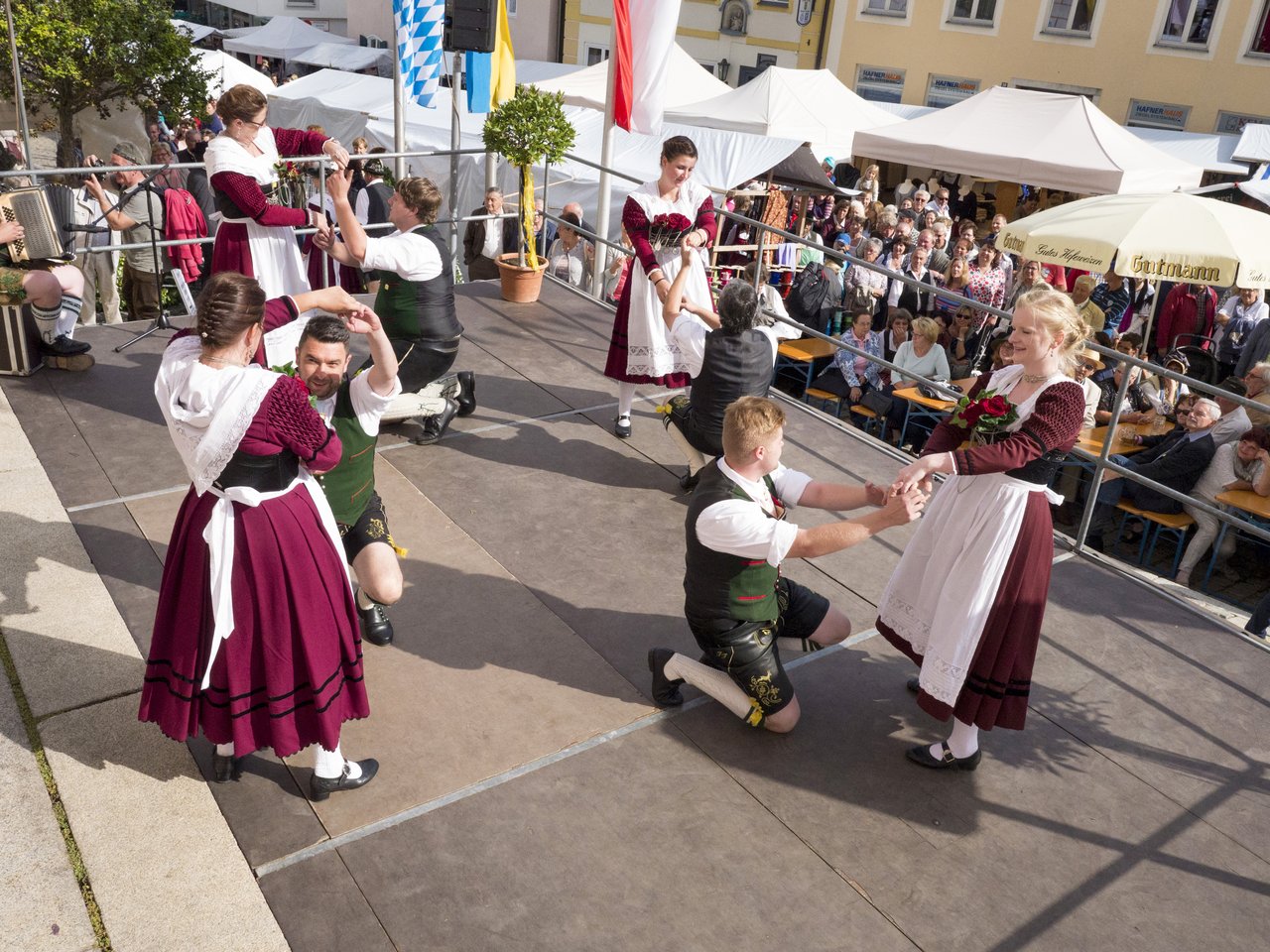 Tanzende Leute in traditionellen Trachten tanzend auf einer Bühne. Das Publikum im Hintergrund staunt.