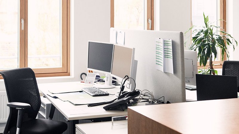 Symbolbild: Ein Blick in ein leeres Büro. Es ist ein Schreibtisch zu sehen, auf dem ein PC-Bildschirm und ein Telefon steht. Davor steht ein Schreibtischstuhl. Im Hintergrund befinden sich große Fenster.