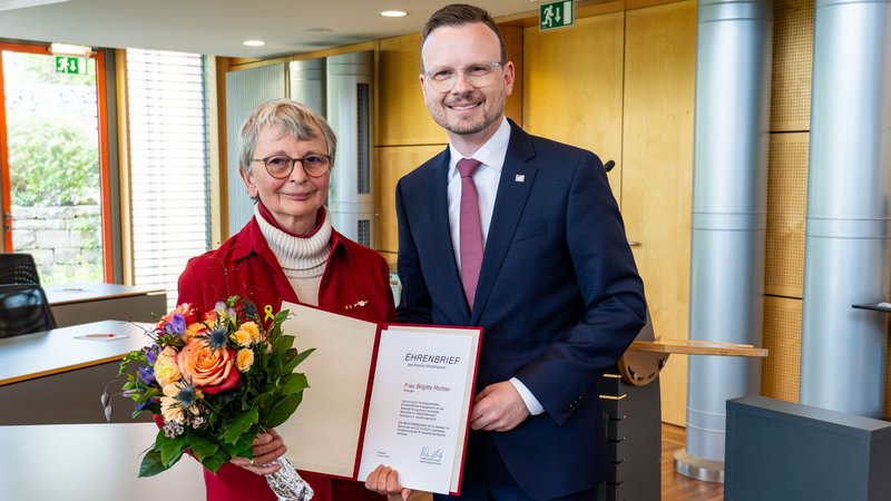 Brigitte Richter und Bezirkstagspräsident Peter Daniel Forster lachen für ein Foto in die Kamera. Brigitte Richter hält dabei den verliehenen Ehrenbrief und einen Blumenstrauß in ihrer Hand.