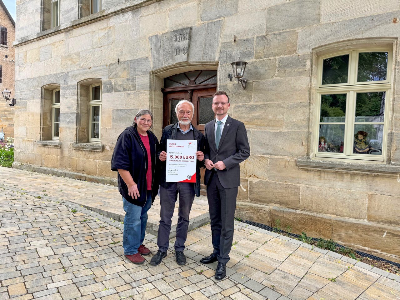 In Henfenfeld überreicht Bezirkstagspräsident Peter Daniel Forster den Förderbescheid über 15.000 Euro an Helmut Wendler. Die beiden stehen zusammen mit einer Frau vor dem sanierten Haus und halten den Förderbescheid in die Kamera.