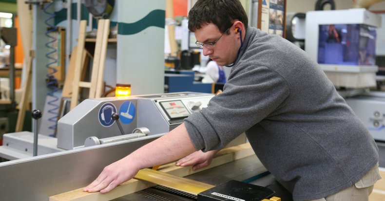 Ein Mann steht in einer großen Fertigungshalle an einem Gerät und schneidet eine Holzlatte.