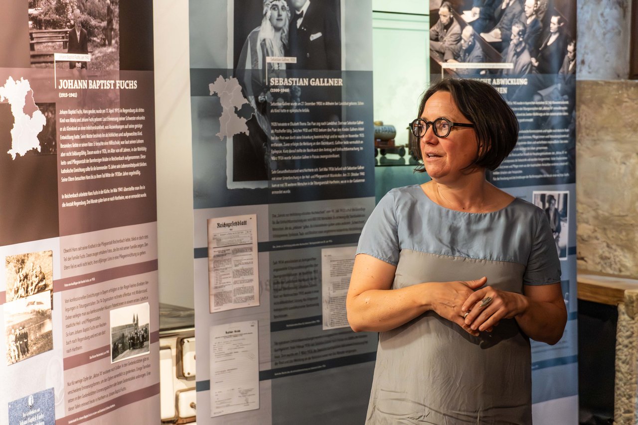 Katrin Kasparek von der Bezirksheimatpflege des Bezirks Mittelfranken steht erklärend vor Rollups der Ausstellung „Ermordet in Grafeneck, Hartheim und Pirna-Sonnenstein – Die Opfer der ,Aktion T4‘ aus Bayern“.