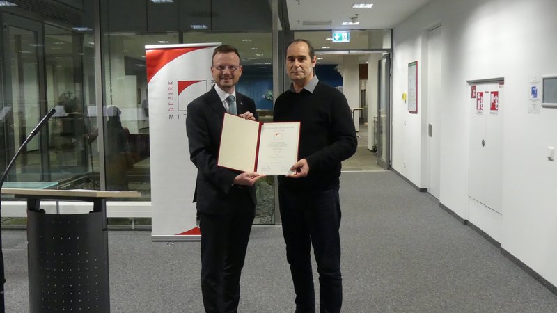 Bezirkstagspräsident Peter Daniel Forster und Preisträger 2024 Timo Reger halten für ein Foto zusammen die Urkunde hoch und lachen in die Kamera.