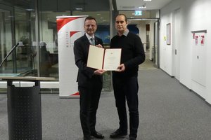 Bezirkstagspräsident Peter Daniel Forster und Preisträger 2024 Timo Reger halten für ein Foto zusammen die Urkunde hoch und lachen in die Kamera.