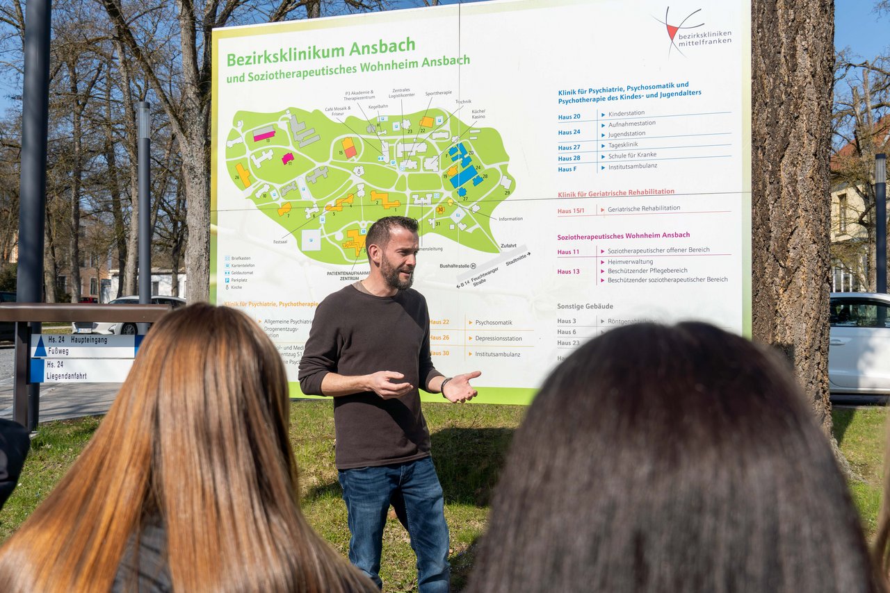 Benjamin Fischer steht vor einer Tafel, auf der der Geländeplan der Bezirkskliniken abgebildet ist und spricht zu den Schülern.