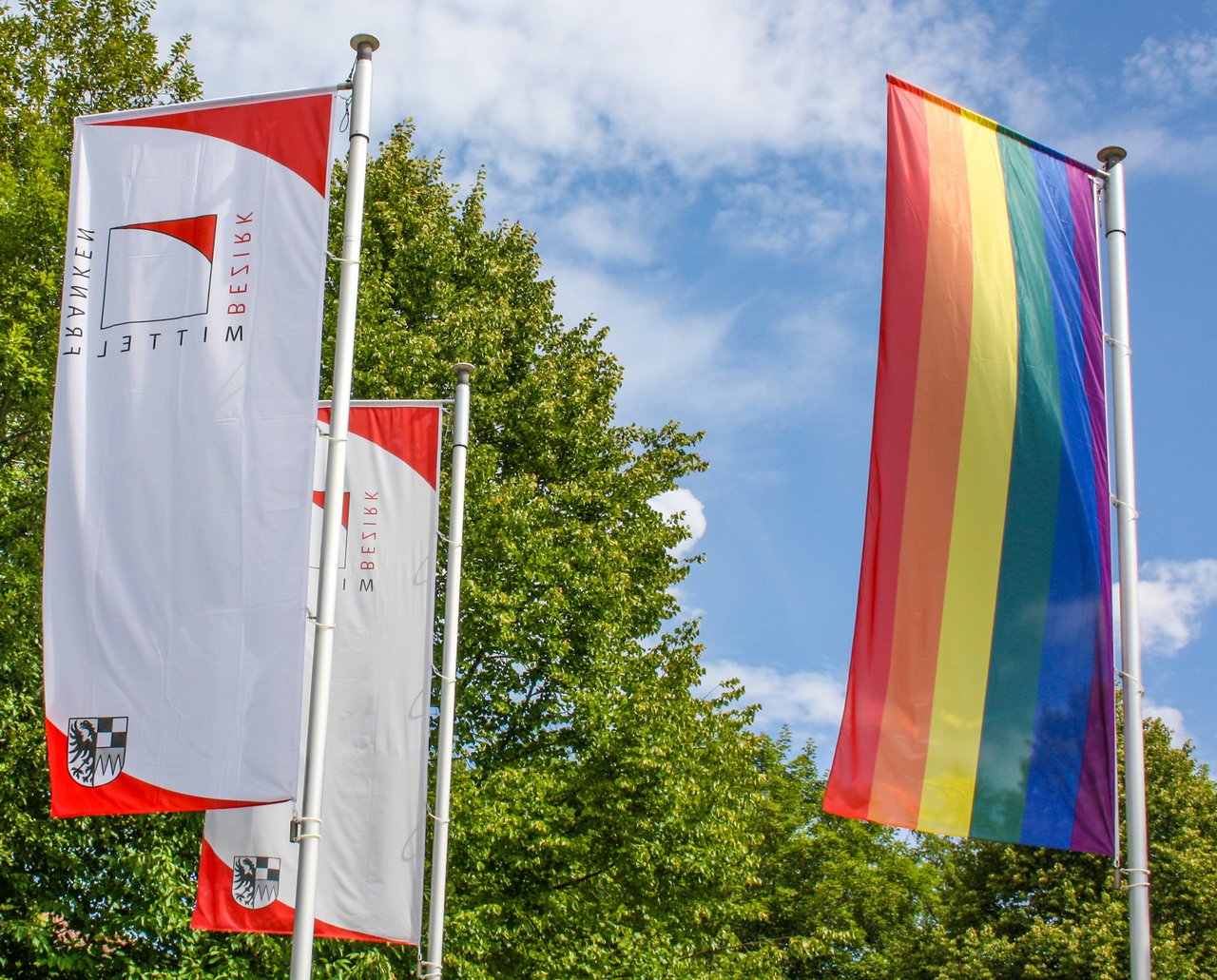 Foto der wehenden Regenbogenfahne am Fahnenmast neben zwei Bezirks-Fahnen.