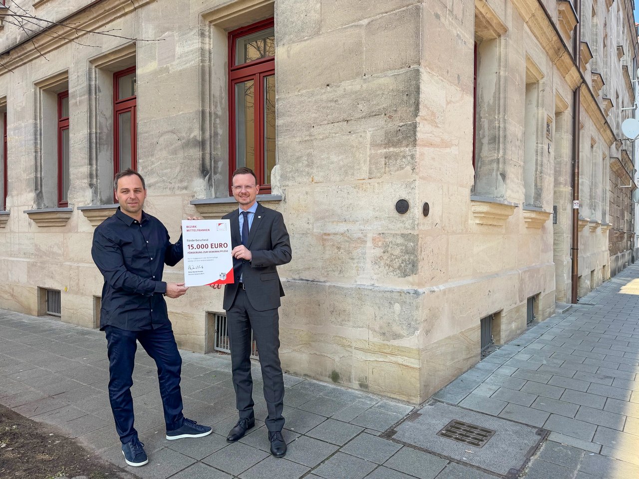 Bezirkstagspräsident Peter Daniel Forster überreicht den Förderbescheid. Die beiden stehen vor dem sanierten Gebäude.