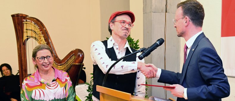 Bezirkstagspräsident Peter Daniel Forster schüttelt einem Künstler am Podium gerade bei der Preisübergabe die Hand.