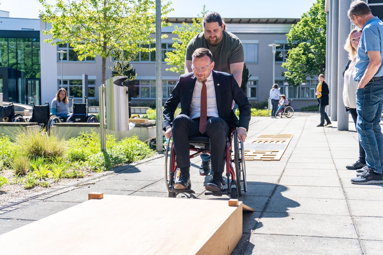 Bezirkstagspräsident Peter Daniel Forster sitzt in einem Rollstuhl und versucht gerade, im Rollstuhlparcours auf eine Rampe hinaufzufahren. Dabei wird er von einem Mann unterstützt, der hinter ihm den Rollstuhl hält und leicht mitschiebt.