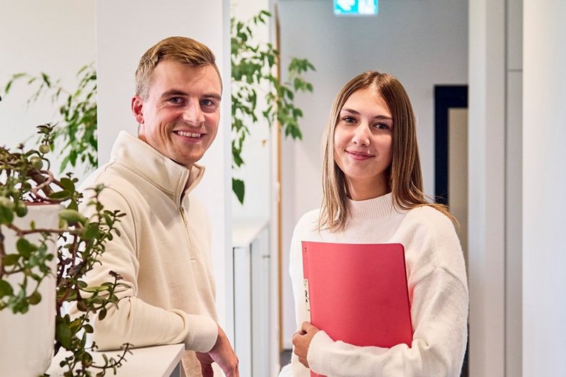 Zwei Personen stehen im Gang eines Büros und schauen in die Kamera.
