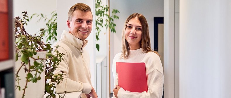 Zwei Personen stehen im Gang eines Büros und schauen in die Kamera.