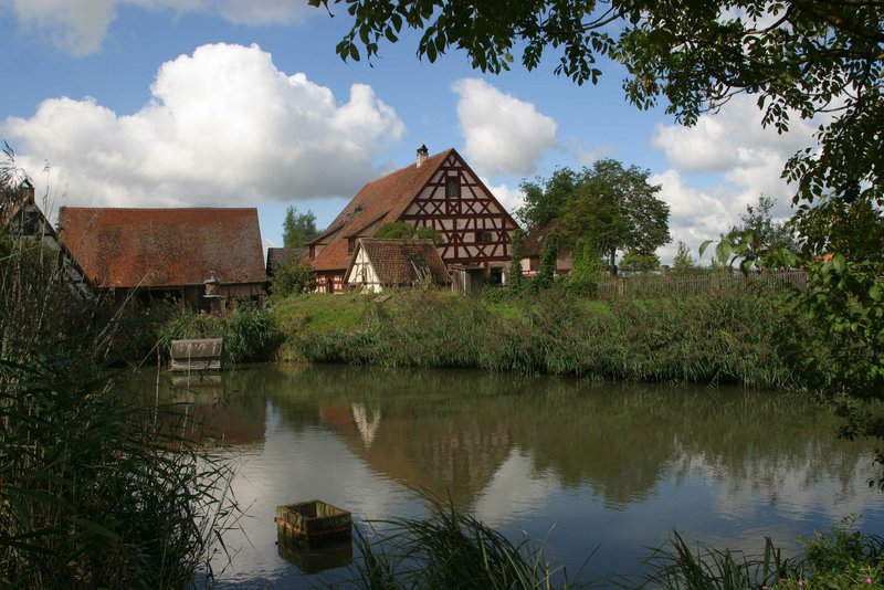 Blick über einen Weiher. Dahinter steht ein altes Fachwerkhaus mit einer Scheune.