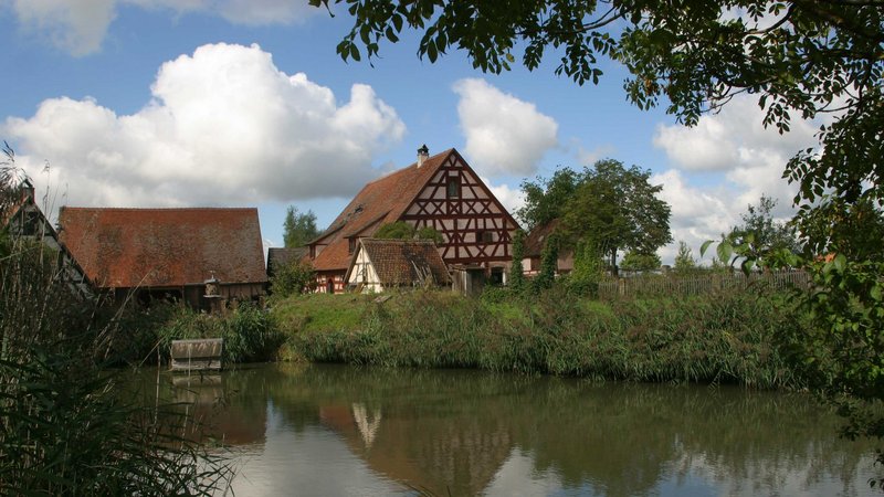 Blick über einen Weiher. Dahinter steht ein altes Fachwerkhaus mit einer Scheune.