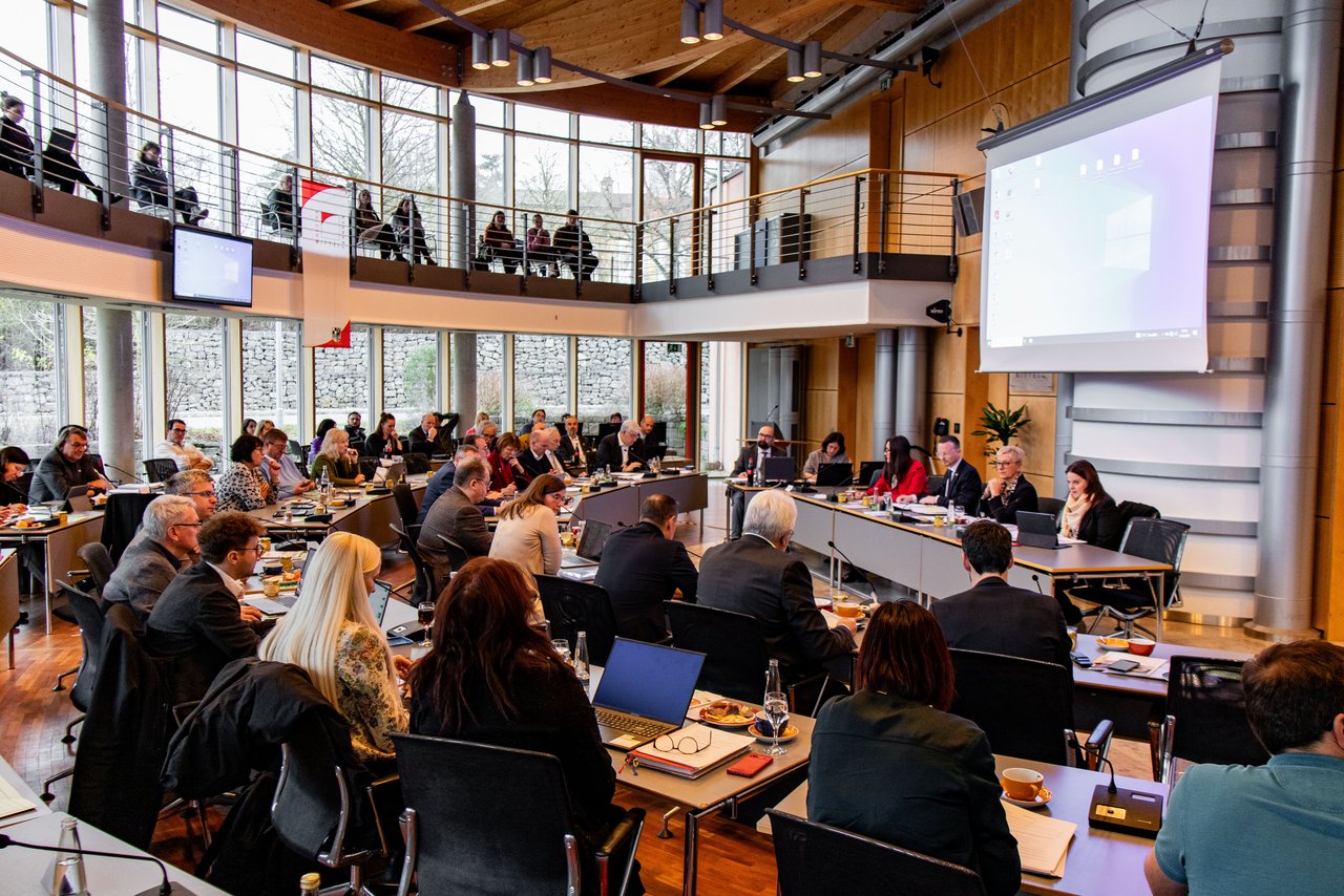 Bild vom Sitzungssaal während einer Sitzung des Bezirkstags. Die Reihen sind voller Menschen, die nach vorne zur Sitzungsleitung schauen.