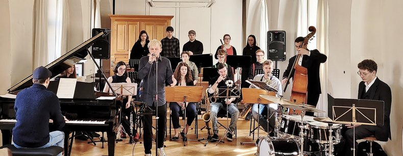 Junge Musikerinnen und Musiker in schwarzen Anzügen machen in festlicher Kulisse gemeinsam Musik.