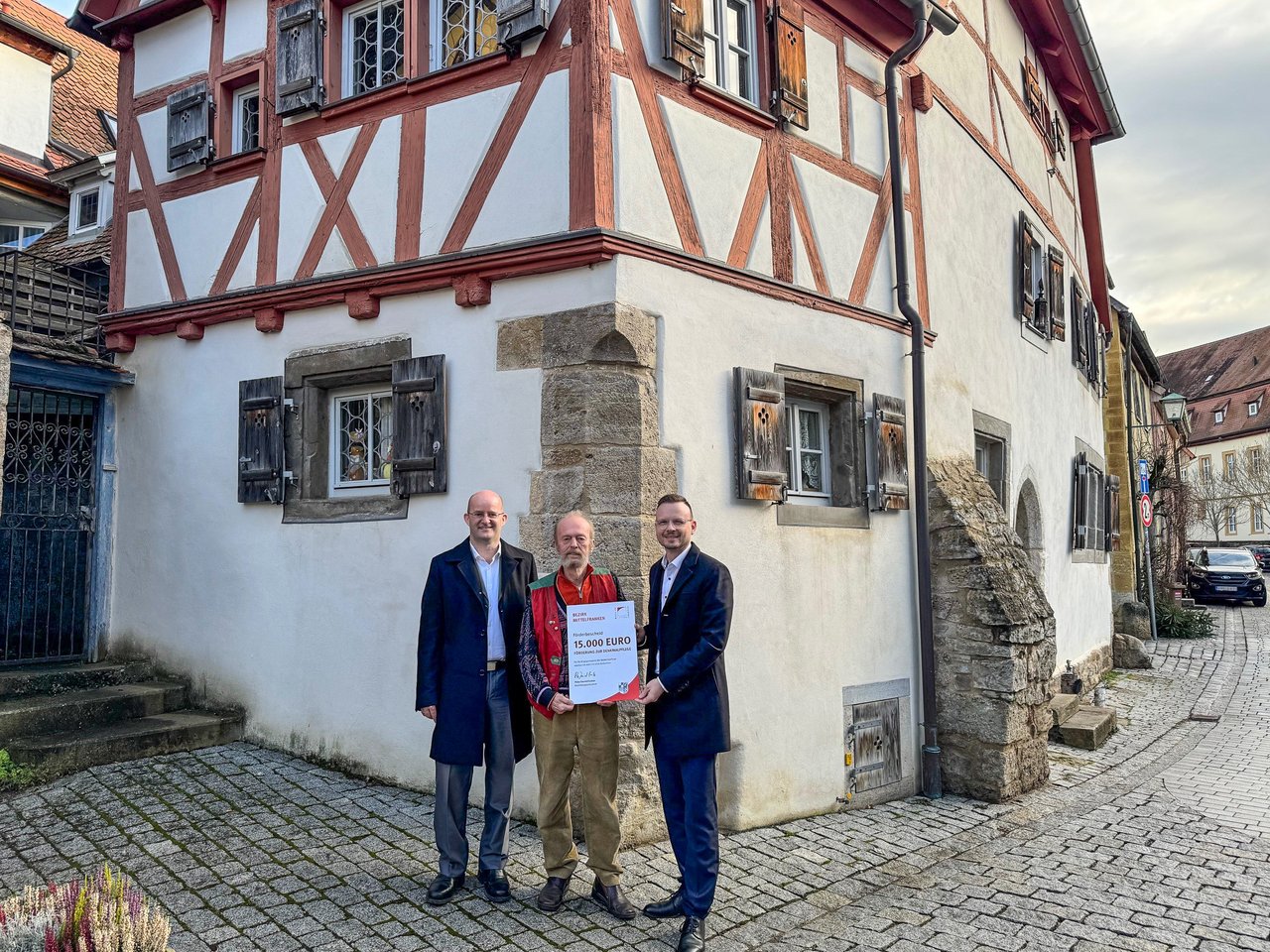 Bezirkstagspräsident Peter Daniel Forster überreicht den Förderbescheid im Beisein des Oberbürgermeisters von Rothenburg o.d.T. Dr. Markus Naser. Sie stehen vor dem sanierten Gebäude.