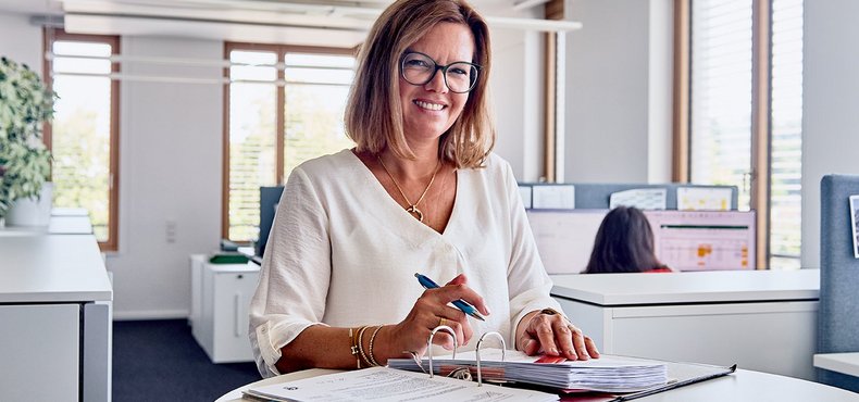 Eine Frau steht an einem Tisch mit einem Stift in der Hand und lacht in die Kamera. Vor ihr auf dem Tisch liegt ein aufgeschlagener Ordner, an dem sie arbeitet.