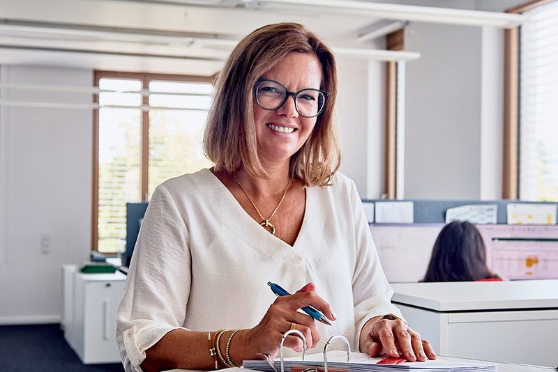 Eine Frau steht an einem Tisch mit einem Stift in der Hand und lacht in die Kamera. Vor ihr auf dem Tisch liegt ein aufgeschlagener Ordner, an dem sie arbeitet.