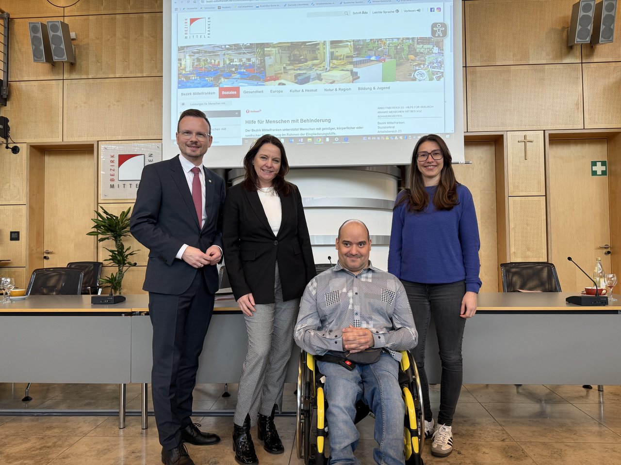 Bezirkstagspräsident Peter Daniel Forster, Regierungspräsidentin Dr. Kerstin Engelhardt-Blum, Metin Ürentoyun und Lisa Tesmer vom Sozialdienst haben sich für ein Gruppenfoto im Sitzungssaal des Bezirksrathauses aufgestellt.