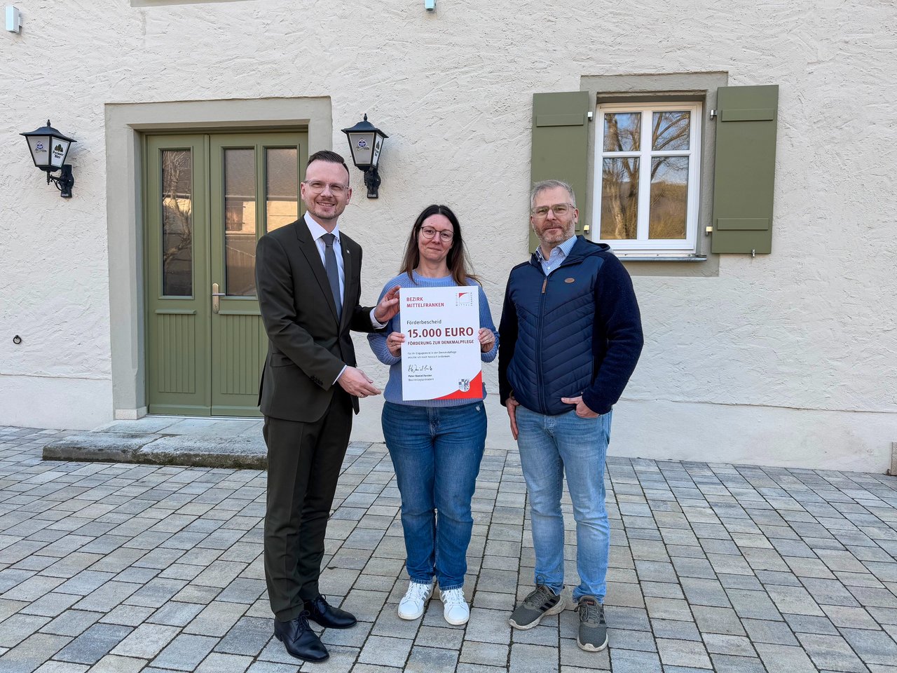 Bezirkstagspräsident Peter Daniel Forster überreicht den Förderbescheid in Weißenburg an Silke und Robert Loy. Sie stehen vor dem sanierten Gebäude.
