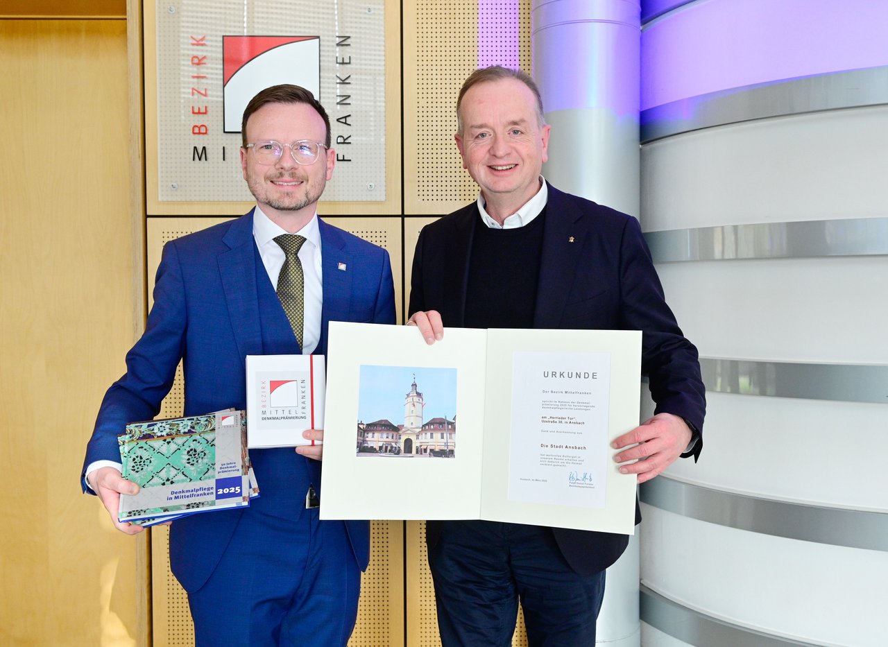 Bezirkstagspräsident Forster und Ansbachs Oberbürgerbeister Deffner stehen nebeneinander und halten die übberreichten Sachen in die Höhe.