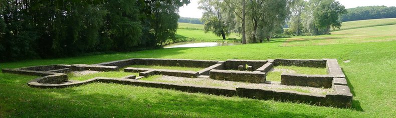 Die Umrisse eines alten Römischen Gebäudes im Boden einer Wiese zu sehen. Drum herum befinden sich Bäume und ein Weiher im Hintergrund.